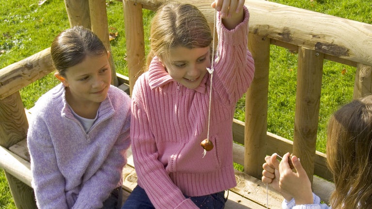 Three young girls sitting on a bench outside holding conkers on strings